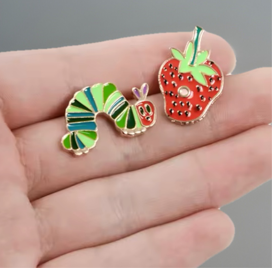 Colorful caterpillar enamel pins and red strawberry pin held in hand by Reed’s Rubber Ducks and Sensory Shop