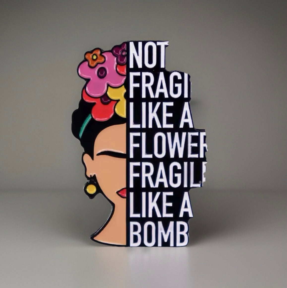 Frida inspired pin featuring half face with colorful flowers and bold text saying not fragile like a flower fragile like a bomb