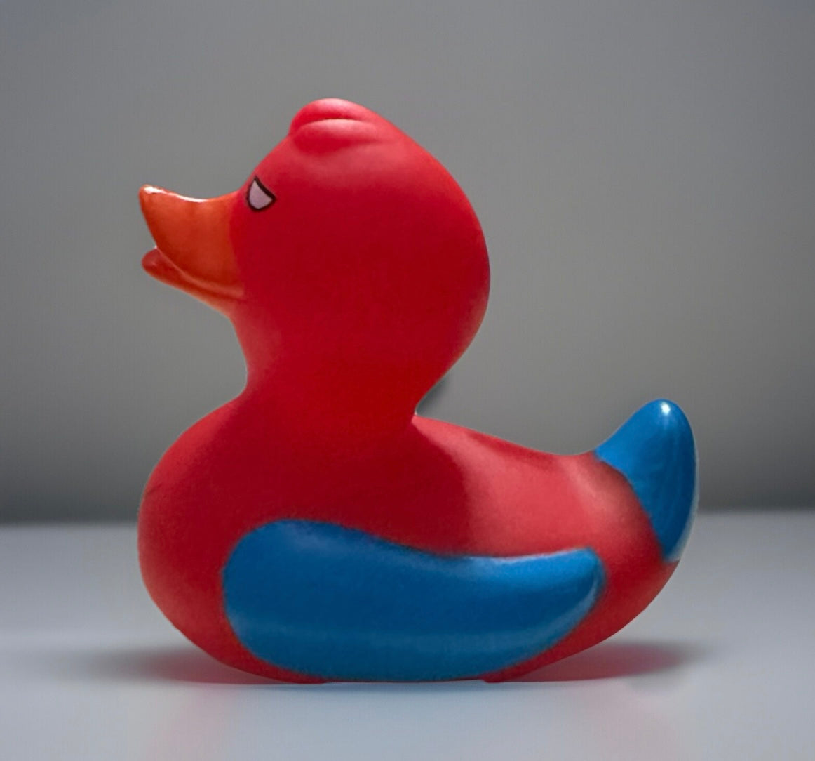 Red and blue Spider Man rubber duck with orange beak and stylized eyes sitting on a gray surface
