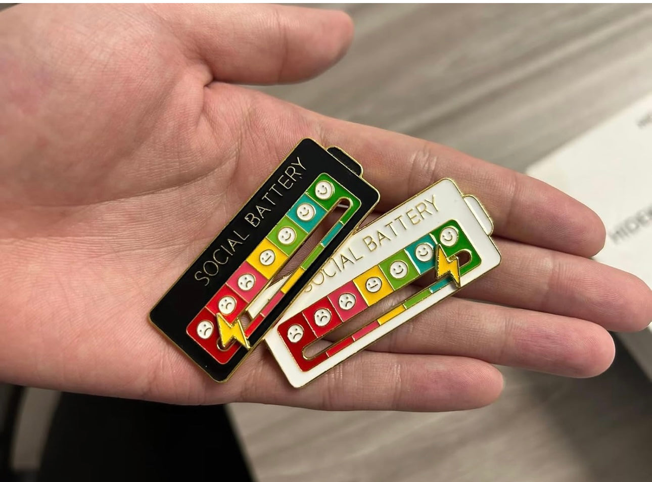 Hand holding two social battery enamel pins one black and one white lapel pin with battery level and emojis
