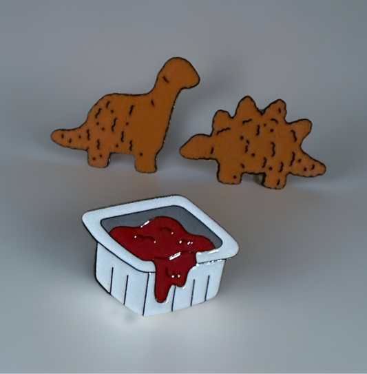 Dinosaur nugget enamel pin set featuring two orange dinosaur shapes and a white dipping sauce container pin